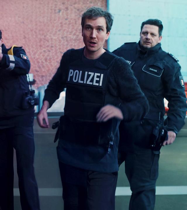 Simon (Jan Krauter) im Einsatz mit schusssicherer Polizeiweste. Hinter ihm zwei Kollegen.