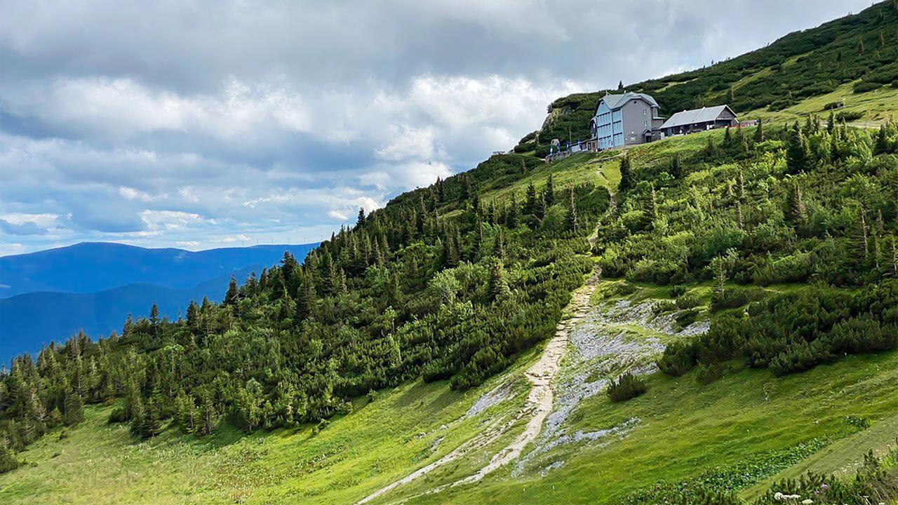 Das Bild zeigt eine hügelige Landschaft in den Wiener Alpen. Im Vordergrund verläuft ein schmaler, gewundener Pfad, der aus hellem, sandigem Erde besteht und sanft den Hang hinauf führt. Auf der linken Seite sind grüne, dichte Sträucher und kleine Bäume zu sehen, die für die alpine Flora typisch sind.  Im Hintergrund steht ein größeres Gebäude mit einer blauen Fassade und einem spitzen Dach. Daneben befinden sich mehrere kleinere Gebäude, die alle umgeben sind von der üppigen Vegetation der Berglandschaft. Der Himmel ist leicht bewölkt mit grauen Wolken, es gibt jedoch auch einige Aufhellungen.  In der Ferne kann man die schemenhaften Umrisse weiterer Berge erkennen, die den Horizont bilden. Das gesamte Bild vermittelt eine friedliche, natürliche Atmosphäre, die typisch für Urlaubsregionen in den Alpen ist.