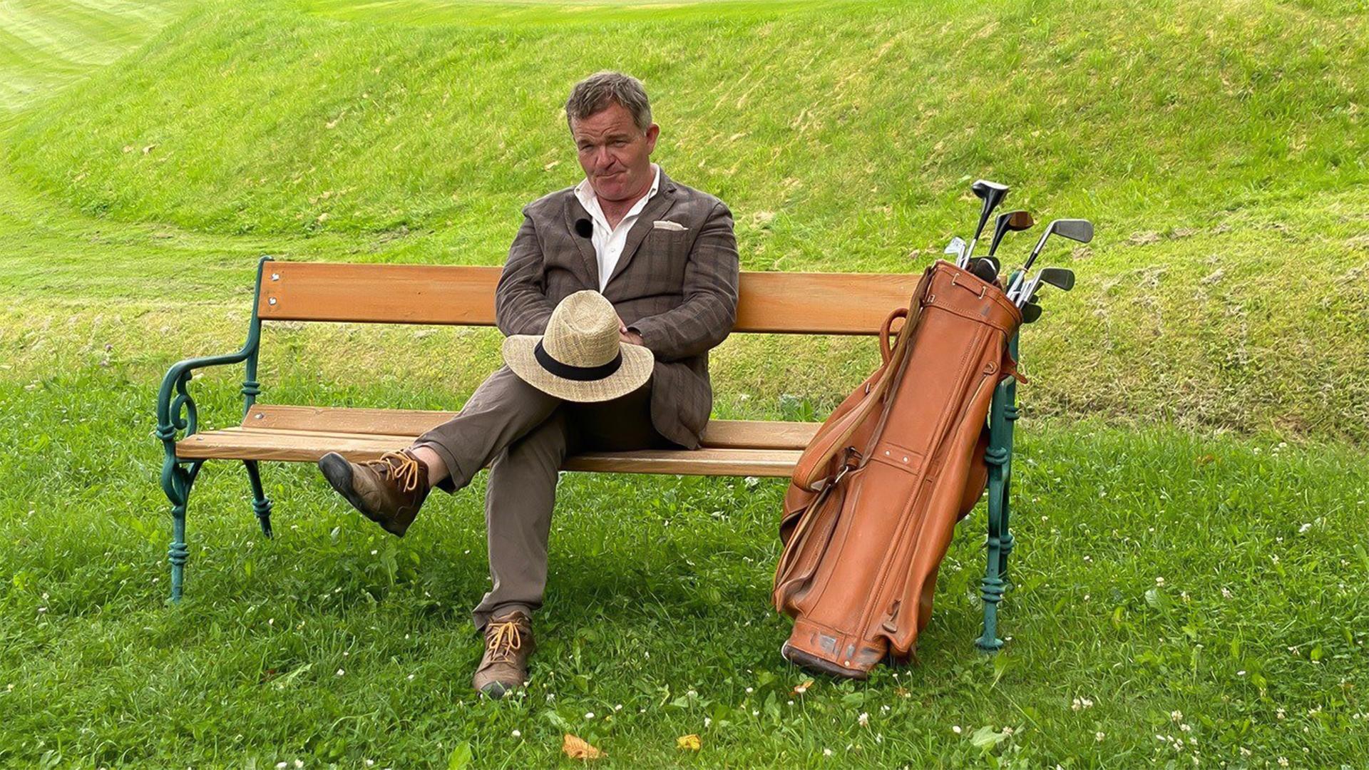 Das Bild zeigt einen Mann, der auf einer Bank sitzt. Er trägt ein kariertes Jackett, ein Hemd und eine Hose. Auf seinen Knien hält er einen Strohhut, der mit einem schwarzen Band verziert ist. Neben ihm steht eine Golf-Tasche aus Leder, aus der mehrere Golfschläger herausragen. Im Hintergrund ist eine grüne Rasenfläche zu sehen, die sanfte Hügel aufweist. Das gesamte Szenario vermittelt eine entspannte, sommerliche Atmosphäre.