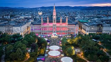 Das Bild zeigt den Wiener Rathausplatz während des Eurovision Villages. Im Vordergrund ist eine große Menschenmenge zu sehen, die vor einem künstlerisch gestalteten Containergebäude steht, das von bunten Lichtern erleuchtet wird. Am Boden sind mehrere große, runde Zeltkonstruktionen sichtbar, die ebenfalls mit Lichtern beleuchtet sind.   Im Hintergrund dominiert das imposante Wiener Rathaus, dessen Fassade in verschiedenen Farben angestrahlt wird, vor allem in Pink und Lila. Die Architektur des Rathauses ist gotisch und prächtig, mit vielen Türmchen und Verzierungen. Hinter den Gebäuden sind sanfte Hügel zu erkennen, die die Silhouette der Stadt ergänzen. Der Himmel ist in sanften Blau- und Grautönen gehalten, was auf die Abenddämmerung hinweist.   Im gesamten Bild sind reichlich Bäume und Grünflächen vorhanden, die dem Platz eine lebendige und einladende Atmosphäre verleihen. Die gesamte Szene vermittelt den Eindruck eines festlichen, geselligen Ereignisses.