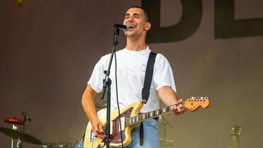 Ein junger Mann steht auf einer Bühne vor einem Mikrofon. Er singt und lacht ins Mikro und spielt dabei auf einer Gitarre, die er umhängen hat.