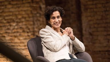 Eine Frau sitzt auf einem grauen Sessel und verschränkt die Hände.