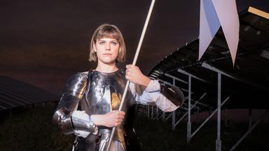 Johanna (Llewellyn Reichman) steht in Rüstung auf einem Feld mit Solarpaneelen. Sie trägt einen weißen Gambeson unter silbernen Metallplatten und hält eine weiße Fahne mit beiden Händen. Hinter ihr ist die Konstruktion der Solaranlage erkennbar, der Himmel wirkt dunkel und dramatisch. 
