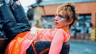 Das Bild zeigt eine Person, die lässig auf einem Stapel Reifen liegt. Die Person trägt ein auffälliges, farbenfrohes Outfit mit einem leuchtend pinken Oberteil und einer orange-pinken, durchscheinenden Rock-ähnlichen Schicht. Die Hose ist dunkel mit einem Tarnmuster. Das Gesicht der Person ist markant geschminkt, mit blauen Akzenten um das rechte Auge und roten Lippen. Die Haare sind kurz und wild gestylt, in einem rötlich-blonden Ton. Im Hintergrund ist eine unscharfe städtische Umgebung mit Gebäuden und einem leicht bewölkten Himmel zu sehen.