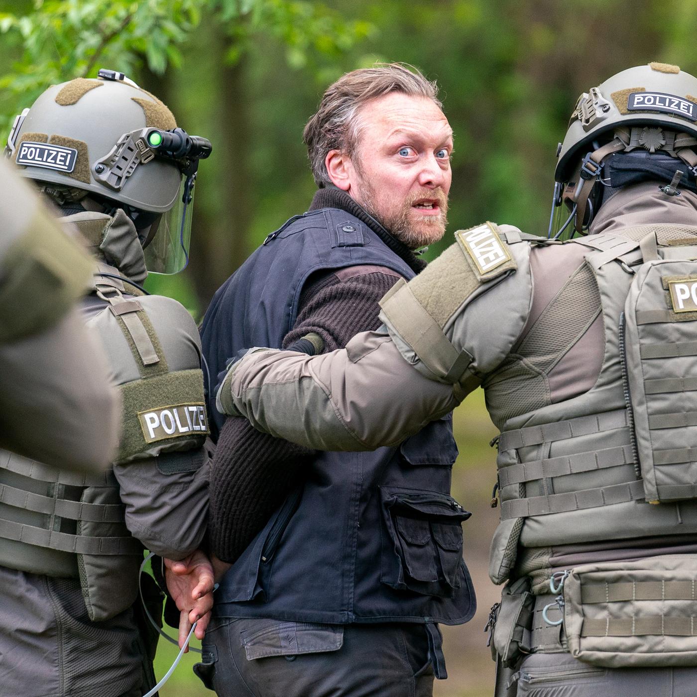 Ein Mann wird im Wald von zwei schwer ausgerüsteten SEK-Beamten der Polizei abgeführt und blickt angespannt über die Schulter.