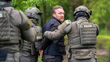 Ein Mann wird im Wald von zwei schwer ausgerüsteten SEK-Beamten der Polizei abgeführt und blickt angespannt über die Schulter.