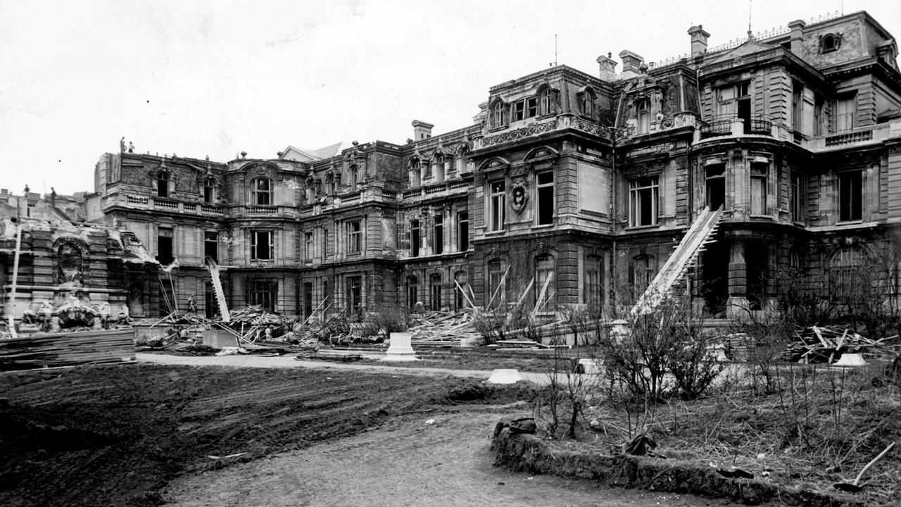 Das Bild zeigt ein stark beschädigtes Palais mit zerbrochenen Fenstern und herausgerissenen Wänden. Baustellenmaterial und Schutt sind verteilt, der Boden ist uneben und erdig.