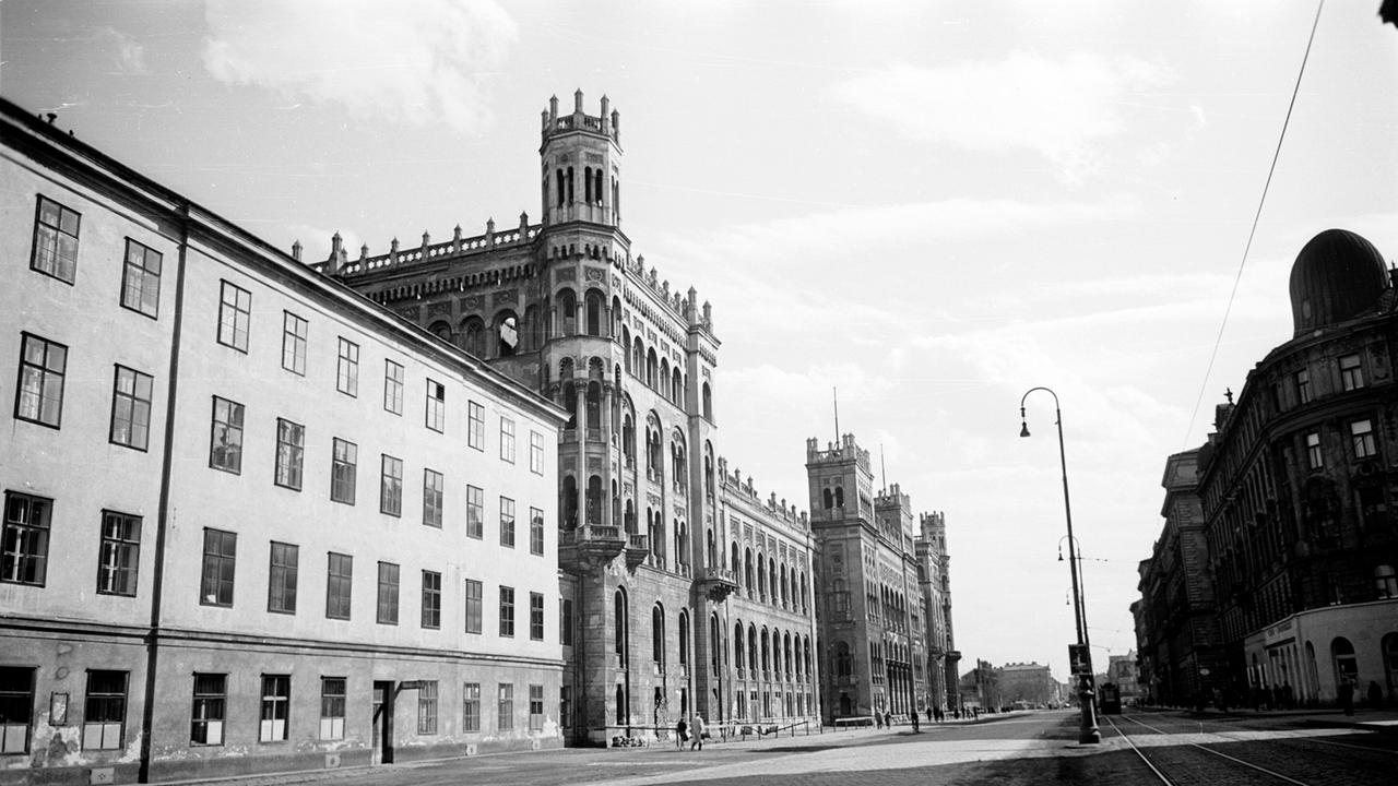 Das Bild zeigt eine Straßenansicht mit historischen Gebäuden in Wien. Links steht ein rechteckiges, mehrstöckiges Gebäude mit vielen Fenstern. Rechts ein aufwendig gestaltetes, gotisches Gebäude mit Türmen. Die Straße ist gepflastert.