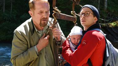 Ein Mann in roter Jacke mit dem Baby auf dem Arm. Und ein Mann der einen Gurt eines Liftes festhält.