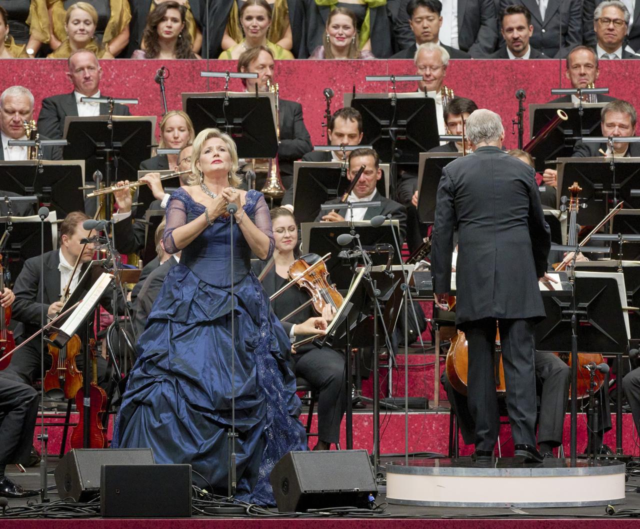 Das Bild zeigt eine Live-Aufführung auf einer Bühne. Im Vordergrund steht eine Sängerin in einem eleganten, dunkelblauen Kleid, das reich mit Falten und einer schulterfreien Gestaltung gearbeitet ist. Sie hat eine nachdenkliche Mimik und hält ein Mikrofon in der Hand. Hinter ihr liegt ein großes Orchester, das aus verschiedenen Musikern besteht, die mit ihren Instrumenten beschäftigt sind. Im Hintergrund sind mehrere Stühle aufgestellt, auf denen Musiker sitzen, die zum Großteil formell gekleidet sind. Die Bühne ist von rotem Samt umgeben, was der Aufführung eine festliche Atmosphäre verleiht.   Im Hintergrund sind auch einige Chormitglieder zu sehen, die in formeller Kleidung gekleidet sind. Es gibt eine Vielzahl von Instrumenten, darunter Geigen, Trompeten und ein Schlagzeug, was auf ein buntes musikalisches Programm hinweist. Der Dirigent, der den Rücken zur Kamera hat, leitet die Musiker. Die gesamte Szene vermittelt das Gefühl eines anspruchsvollen, künstlerischen Events.