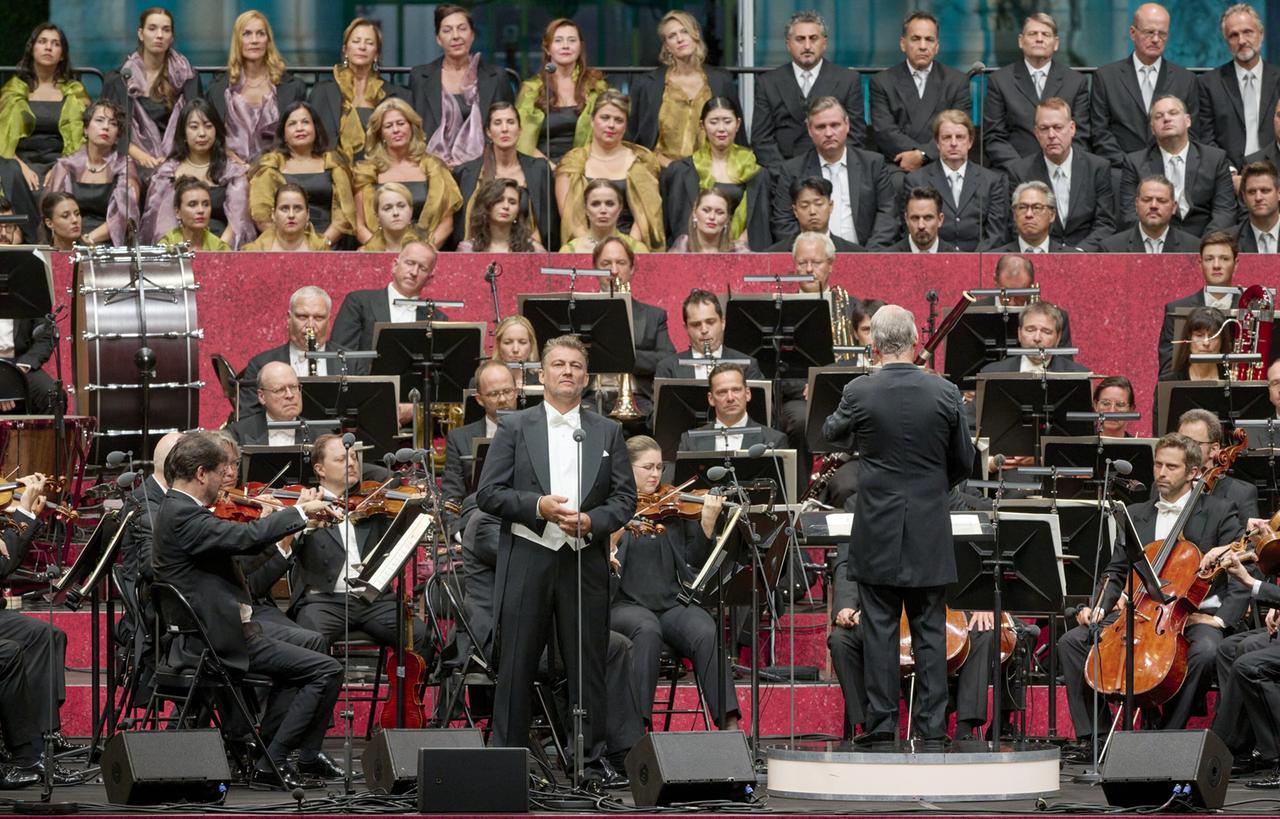 Das Bild zeigt eine Live-Aufführung in einem großen, festlichen Rahmen. Im Vordergrund stehen mehrere Musiker, die auf ihren Stühlen sitzen und ihre Instrumente spielen, hauptsächlich Streichinstrumente. In der Mitte ist ein Sänger zu sehen, der in einem eleganten Anzug auf einem Mikrofon steht und singt. Hinter ihm befindet sich der dirigierende Maestro, der dem Orchester den Takt vorgibt.  Im Hintergrund sieht man einen Chor, der in formellen Outfits gekleidet ist und aufmerksam nach vorne blickt. Die Bühne ist farbenfroh gestaltet, mit pinkfarbenen und roten Elementen, während das Publikum teilweise aus zahlreichen Chormitgliedern besteht.   Die Szenerie vermittelt eine festliche Atmosphäre, die für eine Opernaufführung typisch ist. Die Musiker, Sänger und der Dirigent sind in einem Moment der Darbietung aktiv, was die Energie und Dynamik der Vorstellung widerspiegelt.
