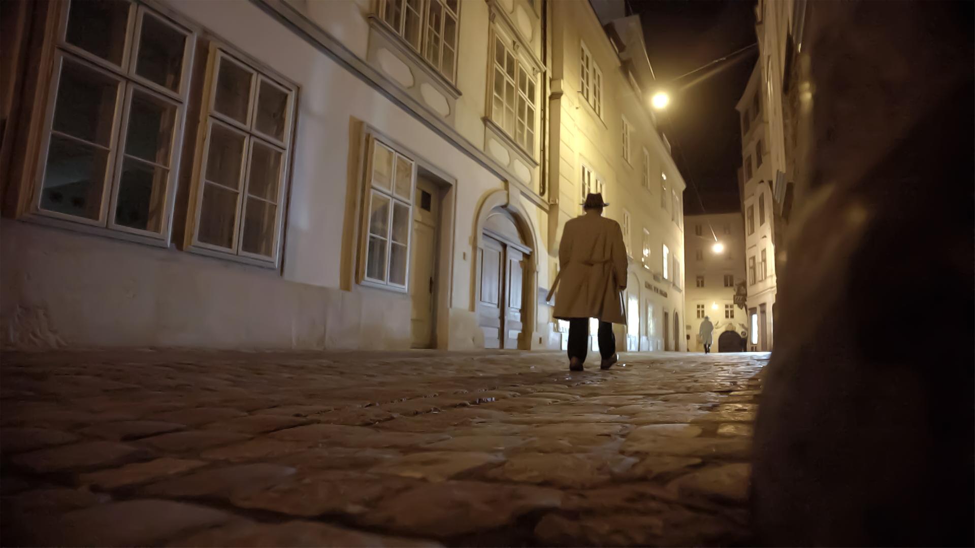 Das Bild zeigt eine nachgestellte Szene aus dem Film „Stadt der Spione“. Es ist eine nächtliche Straßenszene in Wien. Der Gehweg ist aus großen, unregelmäßigen Steinen und wirkt feucht, vermutlich durch Regen. Auf der linken Seite sind alte, historische Gebäude mit großen Fenstern, die durch gelbliche Lichtquellen beleuchtet werden. Die Fassade der Gebäude zeigt eine Mischung aus traditionellen und mehr modernen architektonischen Elementen.  In der Mitte des Bildes geht eine Person mit einem langen, hellen Trenchcoat und einem Hut, die mit dem Rücken zur Kamera steht. Sie bewegt sich auf der Straße in Richtung des oberen Bildrandes. Im Hintergrund ist ein weiteres Gebäude sichtbar, das ebenfalls beleuchtet ist und einige Fenster hat. Die Szene vermittelt ein Gefühl von Geheimnis und Spannung, passend zum Thema Spionage, das der Film behandelt.   Die gesamte Umgebung hat eine düstere, geheimnisvolle Atmosphäre, die durch das Dunkel der Nacht und die gezielte Beleuchtung der Gebäude verstärkt wird.