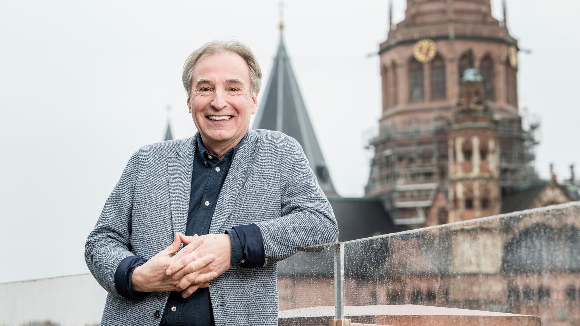 Lächelnder Mann in grauem Tweed-Sakko mit gefalteten Händen, im Hintergrund Turmspitzen einer Kirche.