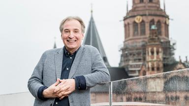 Lächelnder Mann in grauem Tweed-Sakko mit gefalteten Händen, im Hintergrund Turmspitzen einer Kirche.