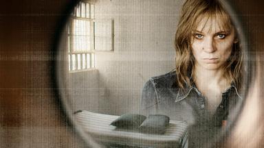 Eine Frau in der Spiegelung einer Glastür. Im Hintergrund eine Zelle.