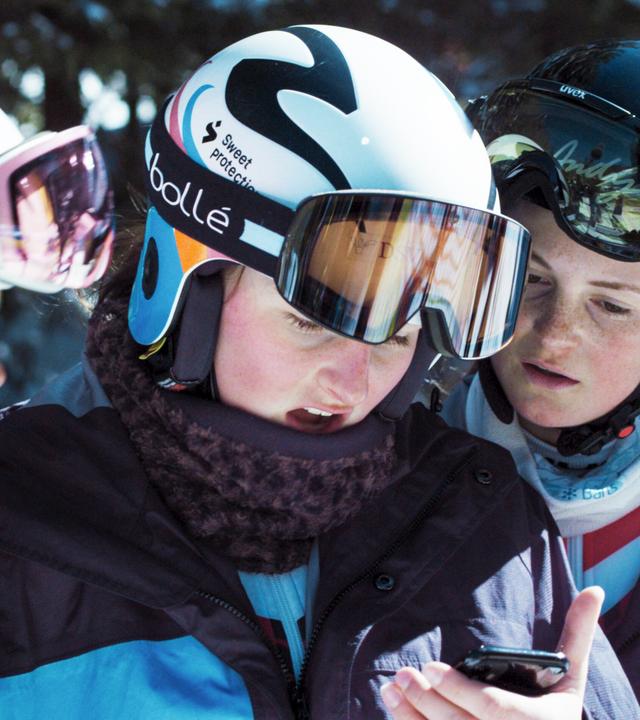 Das Bild zeigt eine Gruppe von drei jungen Skifahrern, die in der Nähe eines Skigebiets sind. Sie tragen Skimützen und Skibrillen. Zwei der Jugendlichen haben Helme auf, einer hat eine farbige Mütze, die bis zu den Ohren reicht. Die Kleidung der Skifahrer ist funktional, mit unterschiedlichen Farbtönen und Skijacken. Sie schauen aufmerksam auf ein Smartphone, während sie offenbar einen Bildschirm oder eine Nachricht darauf betrachten. Im Hintergrund sind unscharfe Konturen von weiteren Personen und schneebedeckten Bäumen zu erkennen. Die Szenerie vermittelt eine winterliche, sportliche Atmosphäre, die auf die engagierte Welt des Skisports hinweist.
