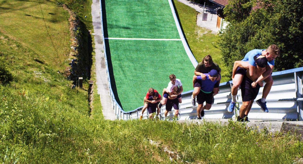 Das Bild zeigt eine Gruppe von jungen Athleten, die auf einer steilen, mit Kunstrasen bedeckten Sportanlage unterwegs sind. Die Sportanlage scheint eine Schanze oder Trainingsstrecke für Skispringen zu sein, mit einem langen, geraden Geländer. Im Vordergrund sieht man mehrere Personen, die in kurzen Hosen und sportlichen Oberteilen gekleidet sind. Einige von ihnen tragen andere Sportler auf ihren Schultern, während sie die Strecke hinaufgehen. Die Umgebung ist grün mit hohem Gras und vereinzelten Bäumen. Im Hintergrund ist ein Gebäude erkennbar, das möglicherweise ein Teil des Sportgeländes ist. Der Himmel ist klar und sonnig. Die Szene vermittelt einen Eindruck von intensivem Training und Teamgeist.