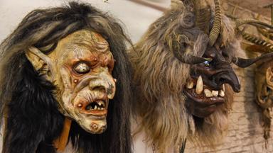 Das Bild zeigt zwei kunstvoll gestaltete Masken, die für den Weihnachtsbrauch in Tirol verwendet werden. Links befindet sich eine Gesichtsmaskerade mit einer gruseligen, froschähnlichen Gestalt, die mit einem dunklen, langen Haaransatz versehen ist. Die Gesichtszüge sind unregelmäßig, mit hervorstehenden Zähnen und hervorgehobenen Augen.  Rechts ist eine weitere Maske zu sehen, die Tiermerkmale aufweist, darunter große Hornstrukturen und ein zotteliges, helles Fell. Diese Maske hat eine breitere Schnauze mit sichtbaren, spitzen Zähnen.  Der Hintergrund des Bildes ist hell und neutrale Wände lassen die Masken hervorstechen, während die maschinenartige und künstlerische Verarbeitung der Masken deutlich erkennbar ist. Diese Masken sind Teil einer Tradition in der Tiroler Gemeinde Stans und repräsentieren den Brauch des "Anklöpfeln".