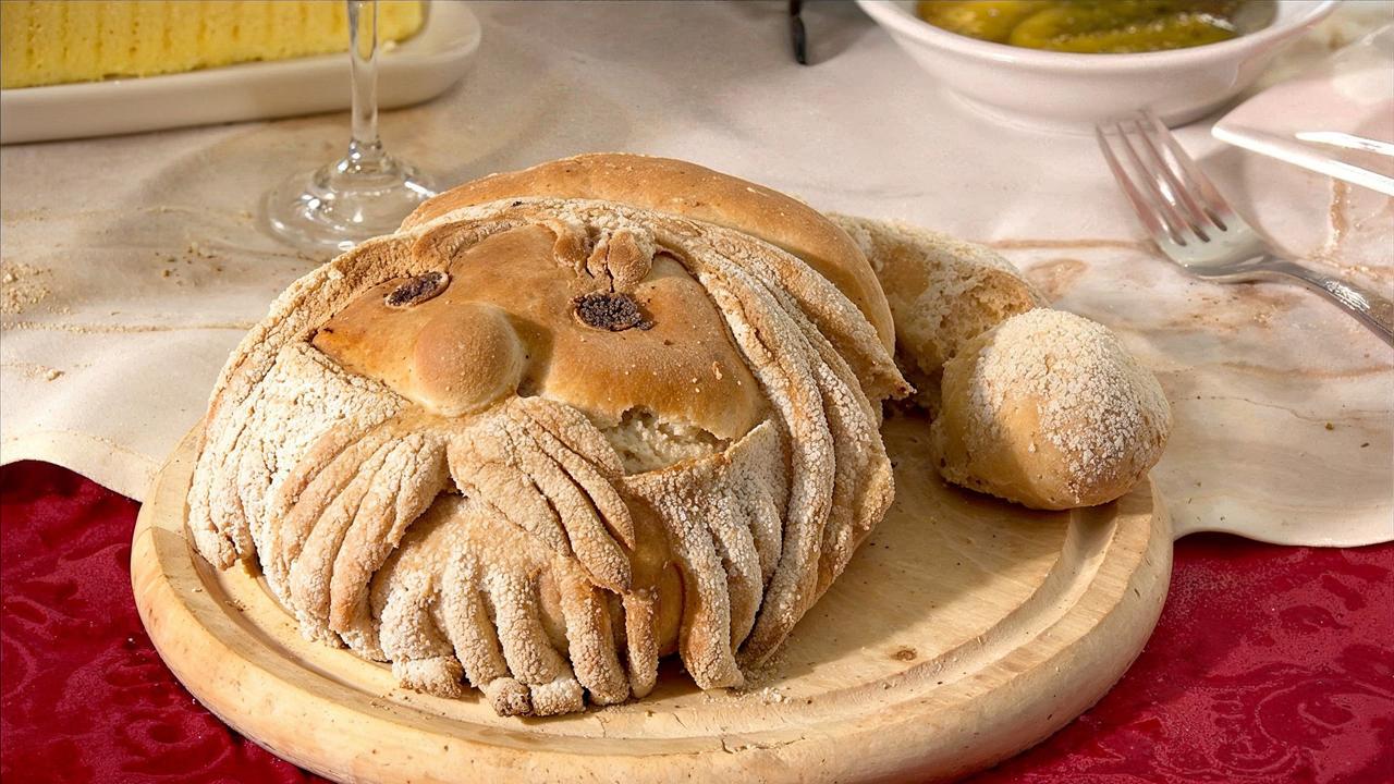 Das Bild zeigt einen liebevoll gestalteten Brotteig in Form eines dicken, bärtigen Mannes, der an den Weihnachtsmann erinnert. Der Brotlaib liegt auf einem runden Holzbrett und hat eine rustikale, krustenartige Oberfläche. Die Gesichtszüge sind mit Rosinen als Augen und einem kleinen runden Stück Teig als Nase dekoriert. Der Bart und die Haare sind mit langen, dünnen Teigsträngen gestaltet, die eine fließende Form erzeugen.   Im Hintergrund ist ein Tisch mit einer roten Tischdecke und weiteren Gegenständen zu sehen, darunter ein Glas, das möglicherweise mit Getränk gefüllt ist, sowie ein weiterer Teller mit unverkennbar anderen Speisen, davon eingelegten Gurken. Dieses Bild vermittelt eine festliche Atmosphäre, die mit Weihnachtsbräuchen und einer traditionellen Mahlzeit in Verbindung steht.