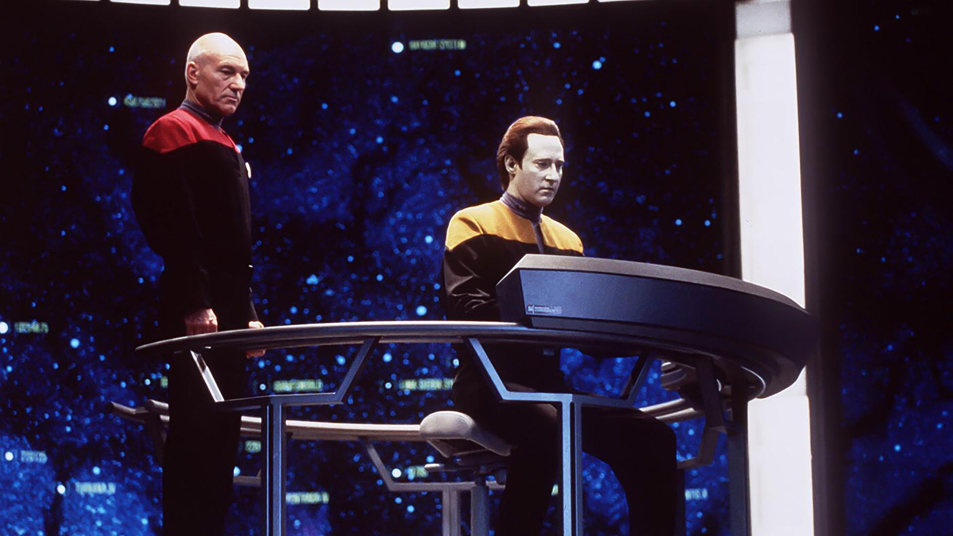 Szene aus der TV-Serie Star Trek Next Generation: Kapitän Picard, ein älterer kahlköpfiger Mann, und der Bordarzt, ein Android, auf der Brücke des Raumschiffs.