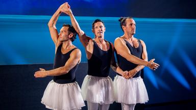 Drei Männer stehen in der Mitte der Bühne und haben Ballettröcke an. Sie halten sich an den Händen. Der Hintergrund ist blau beleuchtet.