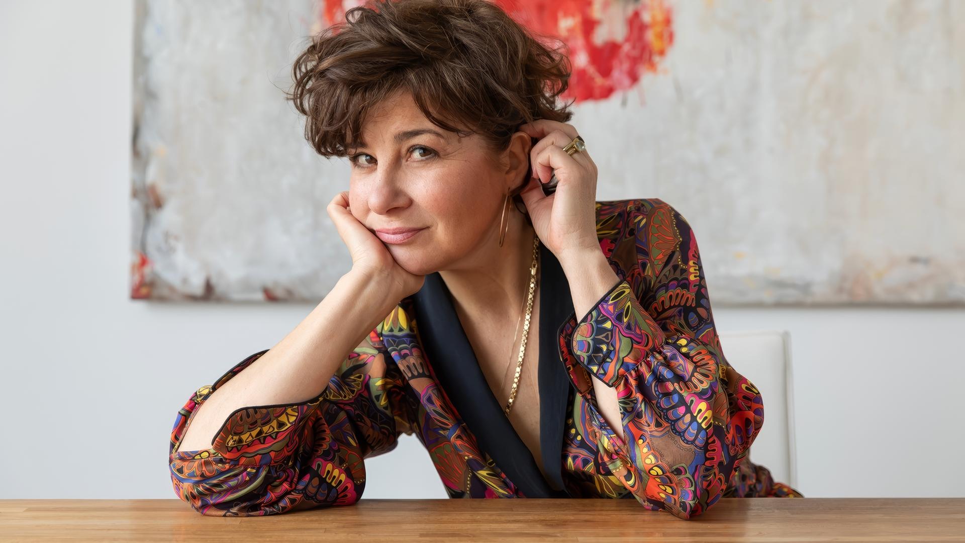 Schauspielerin Anita Vulesica mit kurzem, lockigem Haar, in farbenfroher Bluse, sitzt entspannt an einem Tisch und blickt lächelnd in die Kamera; im Hintergrund ein abstraktes Gemälde.