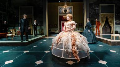 Große Bühnenaufstellung mit einer Frau in einem rosafarbenen, weit ausgestellten Reifrockkleid und roten, gelockten Haaren im Vordergrund; weitere Personen stehen verteilt im Raum eines klassizistischen Salons.