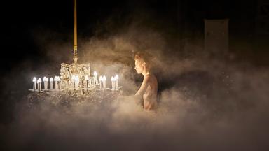 Dichte, weiße Nebelschwaden füllen fast das ganze Bild und geben nur einen kleinen Bereich frei, in dem eine einzelne, schlanke Frau steht. Vor ihr hängt ein großer, prunkvoller Kristalllüster. Der Nebel reflektiert das Licht, sodass eine weiche, flirrende Atmosphäre entsteht. Der Raum dahinter ist dunkel, fast schwarz.