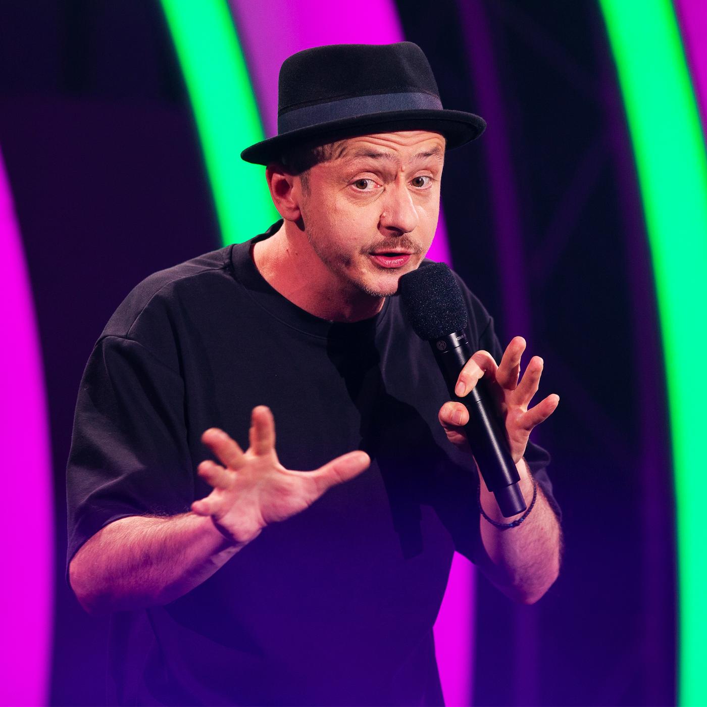 Stefan Danziger steht in der Mitte des Bildes.  In seiner linken Hand hält er ein Mikrofon. Die rechte Hand streckt er mit gespreizten Fingern nach vorne aus. Der Hintergrund ist rosa und grün beleuchtet.