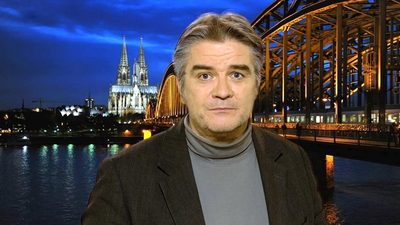 Nahaufnahme von Stefan Koldehoff in einem Studio. Im Hintergrund sieht man den Kölner Dom und die Hohenzollernbrücke.