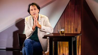 Stefan Waghubinger sitzt in der Mitte des Bildes auf einem Barhocker. Er faltet die Hände. Rechts von ihm steht ein Bartisch. Auf dem Tisch ist ein Bier. Er trägt ein beiges Sakko.