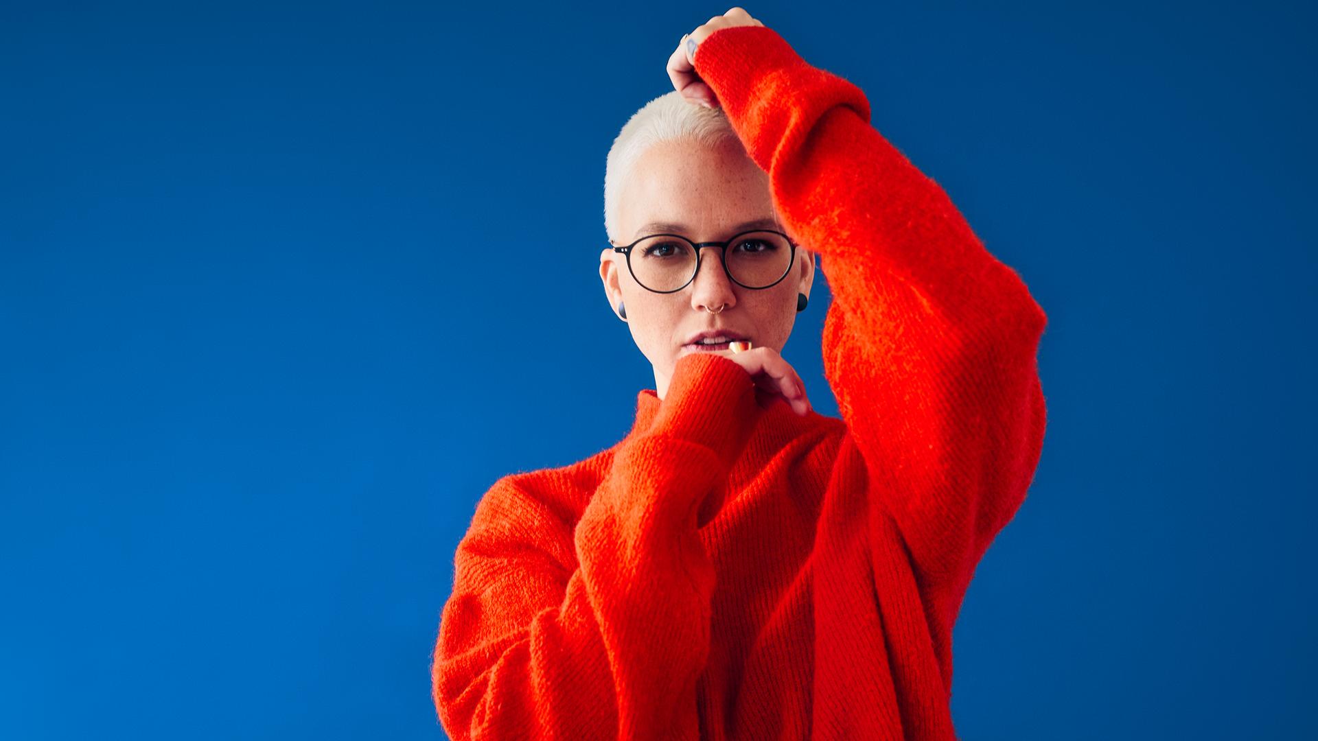 Stefanie Heinzmann steht vor einem blauen Hintergrund. Sie trägt einen roten Pulli und eine Brille. Ihr linker Arm ist schulterhoch gehoben und ihre Hand liegt auf ihrem Kopf. Ihr rechter Arme ist angewinkelt, ihre Handrücken berührt ihr Kinn.