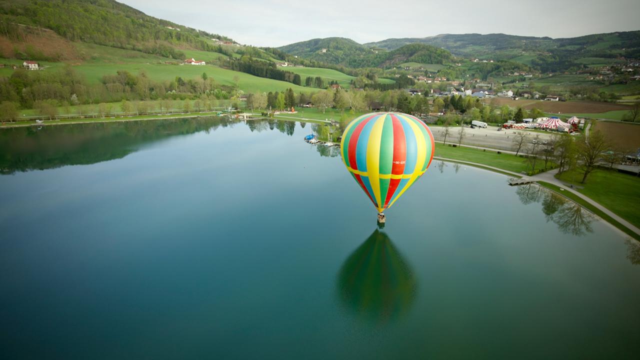 Das Bild zeigt eine malerische Landschaft in der Steiermark, Österreich. Im Vordergrund ist ein Wasseroberfläche zu sehen, die die Umgebung wie einen Spiegel reflektiert. In der Mitte des Bildes schwebt ein bunter Heißluftballon mit rot-gelben und grünen Streifen über dem Wasser.   Um den Ballon herum sind Bäume und Hügel zu erkennen, die mit frischem Grün bedeckt sind und die sanften Konturen der Landschaft betonen. An einem Ufer des Sees sind mehrere kleine Boote zu sehen, die am Steg festgemacht sind. Am anderen Ufer erkennt man einen Park mit Wegen, Bäumen und Wiesen, sowie ein großes, festlich gestreiftes Zelt, das auf eine Veranstaltung hindeutet.   Die gesamte Szene strahlt Ruhe und die natürliche Schönheit der Region aus, die bekannt ist für ihre vielfältigen Landschaften und zahlreiche Seen.