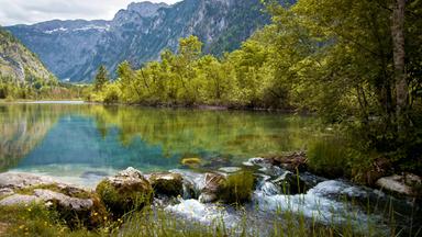 Das Bild zeigt eine idyllische Szenerie im Seenland Österreich, insbesondere im Siebenseengebiet der Steiermark. Im Vordergrund fließt ein kleines, klares Gewässer, das über glatte Steine und zwischen grünen Grasbüscheln hinabplätschert.   Rund um das Wasser wachsen zahlreiche hohe Bäume mit frischem, grünem Laub. Die Vegetation ist üppig und vielfältig. Im Hintergrund erheben sich majestätische Berggipfel, die teilweise mit Schnee bedeckt sind und von Nebel umhüllt werden, was auf wechselhaftes Wetter hindeutet.   Die Wasseroberfläche spiegelt die Umgebung wider und schafft so eine harmonische Verbindung zwischen den verschiedenen Elementen der Natur. Insgesamt vermittelt das Bild eine ruhige und friedliche Atmosphäre, die typisch ist für die Landschaften in der Steiermark.
