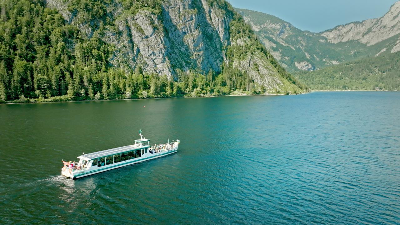 Das Bild zeigt einen ruhigen Gebirgssee in der Steiermark, Österreich, umgeben von hohen, bewaldeten Bergen. Die Wasseroberfläche des Sees ist glitzernd und hat eine blau-grüne Farbe. In der Mitte des Bildes fährt ein Solarschiff, das eine lange, flache Form hat. Das Schiff hat eine transparente Überdachung und scheint mit Passagieren besetzt zu sein. Im Hintergrund sind steile Felsen und dichte Wälder zu sehen, die dem Bild eine natürliche und malerische Kulisse verleihen. Der Himmel ist klar und blau, was eine friedliche Atmosphäre schafft.