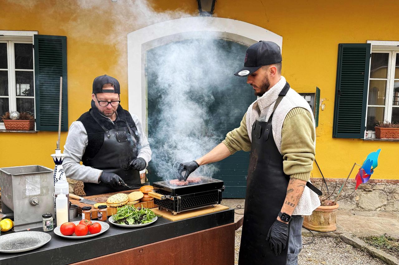 Das Bild zeigt zwei Männer, die an einem Grill stehen. Der Hintergrund besteht aus einem gelben Gebäude mit weißen Fensterrahmen und grünen Fensterläden.   Der Mann links trägt eine schwarze Kappe und eine Brille. Er trägt ein graues Oberteil unter einer schwarzen Schürze und bearbeitet gerade Fleisch. Der Mann rechts hat ebenfalls eine schwarze Kappe, ein helles Sweatshirt mit einem kragenähnlichen Teil aus Fleece und eine schwarze Schürze an. Er ist dabei, ein Stück Fleisch auf dem Grill zu wenden, aus dem Rauch aufsteigt.  Auf dem Tisch vor den Männern sind verschiedene Zutaten angeordnet: Es gibt frische Tomaten, Blattsalat, Ketchup, Gewürze in kleinen Gläsern, sowie Hamburgerbrötchen. In der Nähe des Grills steht eine Grillwanne. Im Hintergrund sind ein paar Töpfe und ein bunter Windspiel sichtbar, das in der Nähe eines Fensters steht.   Die Szenerie vermittelt eine gemütliche Atmosphäre, die mit dem Kochen und der Zubereitung von Speisen verbunden ist.