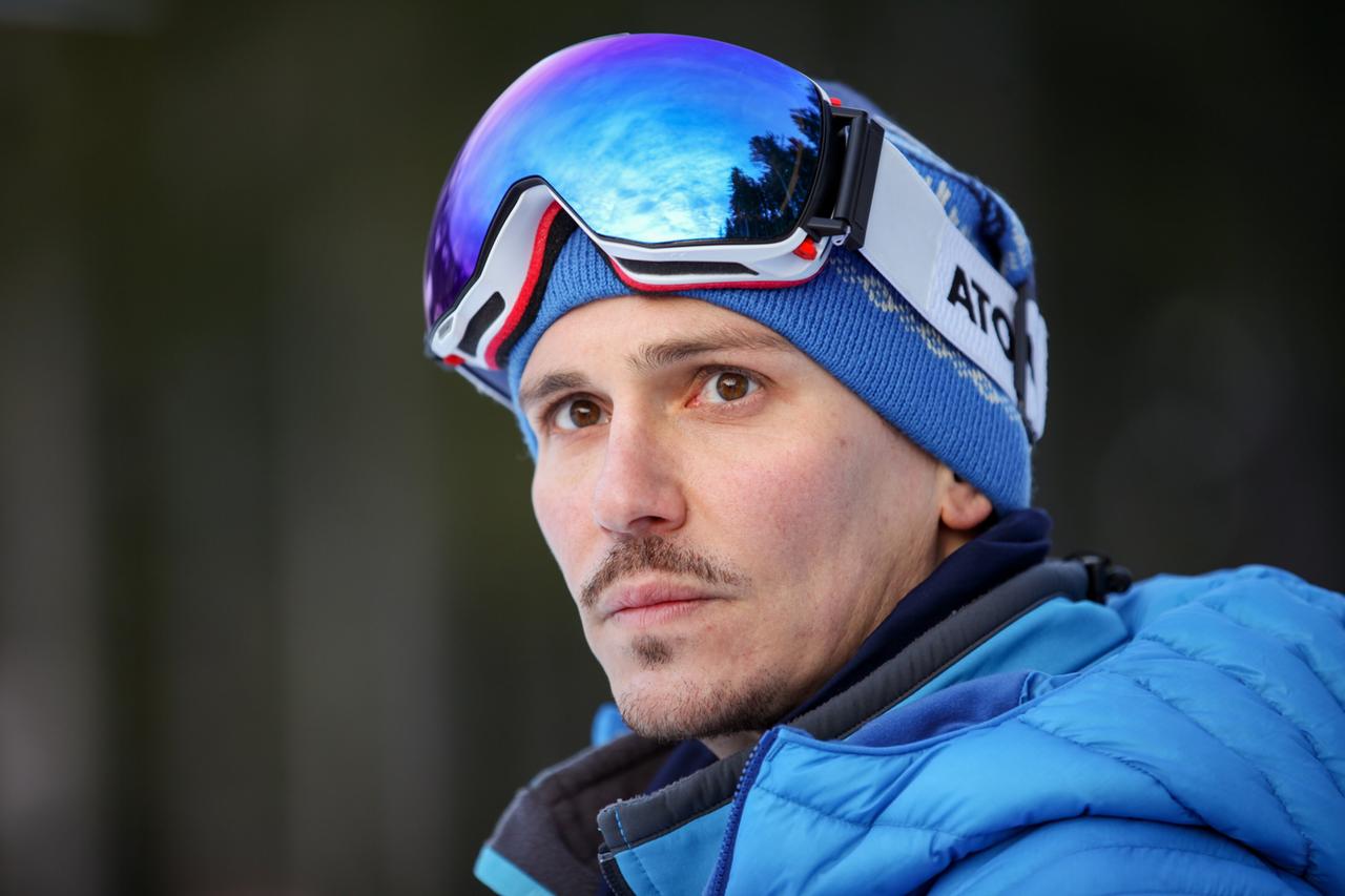 Das Bild zeigt einen Mann, der in den Bergen oder einem geschlossenen Bereich mit Schnee im Hintergrund steht. Er trägt eine blaue Jacke mit einer schwarzen Innenschicht und eine blaue Mütze, auf der der Schriftzug "ATOK" zu sehen ist. Auf seinem Kopf trägt er eine Sonnenbrille mit einer großen, getönten, farbigen Linse, die den Himmel reflektiert. Der Mann hat einen kurzen Bart und schaut direkt in die Kamera, wobei sein Gesicht ernst wirkt. Der Hintergrund ist unscharf und lässt auf eine winterliche Landschaft schließen.