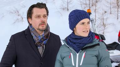 Das Bild zeigt zwei Personen, die in einer winterlichen Umgebung stehen, die von Schnee und Bäumen umgeben ist. Im Hintergrunde sind schneebedeckte Bäume und ein Schneedepot sichtbar.   Die erste Person, ein Mann, hat dunkle, leicht gewellte Haare und einen kurzen Bart. Er trägt einen dunklen Mantel und einen gestreiften Schal. Die zweite Person, eine Frau, hat kurze Haare und trägt eine dunkelblaue Mütze sowie eine graue Winterjacke. Beide scheinen nachdenklich oder besorgt zu sein und schauen in die Ferne. Der Ausdruck auf ihren Gesichtern deutet darauf hin, dass sie in eine ernste Diskussion vertieft sind.   In der Nähe ist ein Schneedepot oder ein Schneemobil erkennbar, was darauf hinweist, dass sich die Szene in einem winterlichen Freizeitgebiet oder einem Skigebiet abspielt.