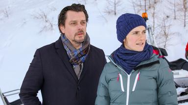 Das Bild zeigt zwei Personen, die in einer winterlichen Umgebung stehen, die von Schnee und Bäumen umgeben ist. Im Hintergrunde sind schneebedeckte Bäume und ein Schneedepot sichtbar.   Die erste Person, ein Mann, hat dunkle, leicht gewellte Haare und einen kurzen Bart. Er trägt einen dunklen Mantel und einen gestreiften Schal. Die zweite Person, eine Frau, hat kurze Haare und trägt eine dunkelblaue Mütze sowie eine graue Winterjacke. Beide scheinen nachdenklich oder besorgt zu sein und schauen in die Ferne. Der Ausdruck auf ihren Gesichtern deutet darauf hin, dass sie in eine ernste Diskussion vertieft sind.   In der Nähe ist ein Schneedepot oder ein Schneemobil erkennbar, was darauf hinweist, dass sich die Szene in einem winterlichen Freizeitgebiet oder einem Skigebiet abspielt.