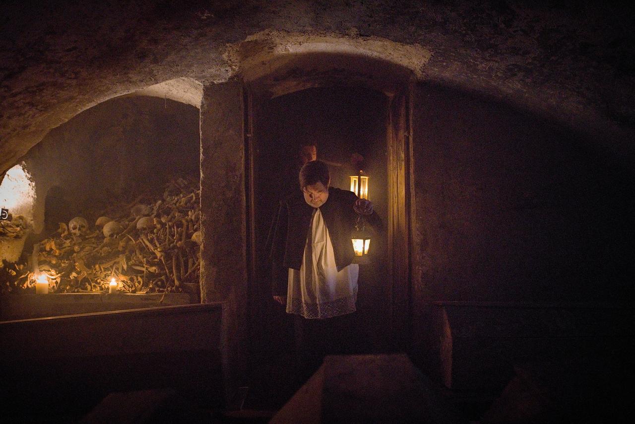 Mann geht durch Kellergewölbe, nebenan gestapelte Gebeine