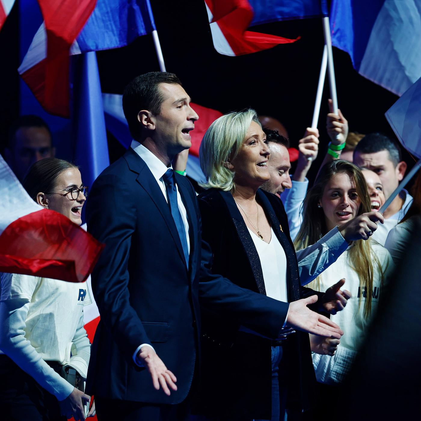Marine Le Pen und Jordan Bardella auf der Bühne, hinter ihnen stehen, mit der französischen Flagge winkend, junge Menschen.