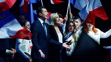 Marine Le Pen und Jordan Bardella auf der Bühne, hinter ihnen stehen, mit der französischen Flagge winkend, junge Menschen.