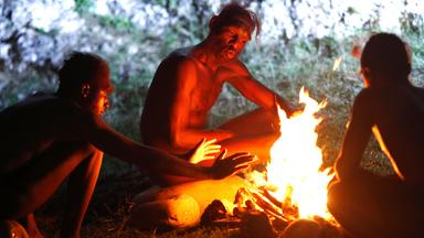 Drei nackte Afrikaner sitzen am Lagerfeuer. Einer von ihnen streckt die Hände in Richtung Feuer.