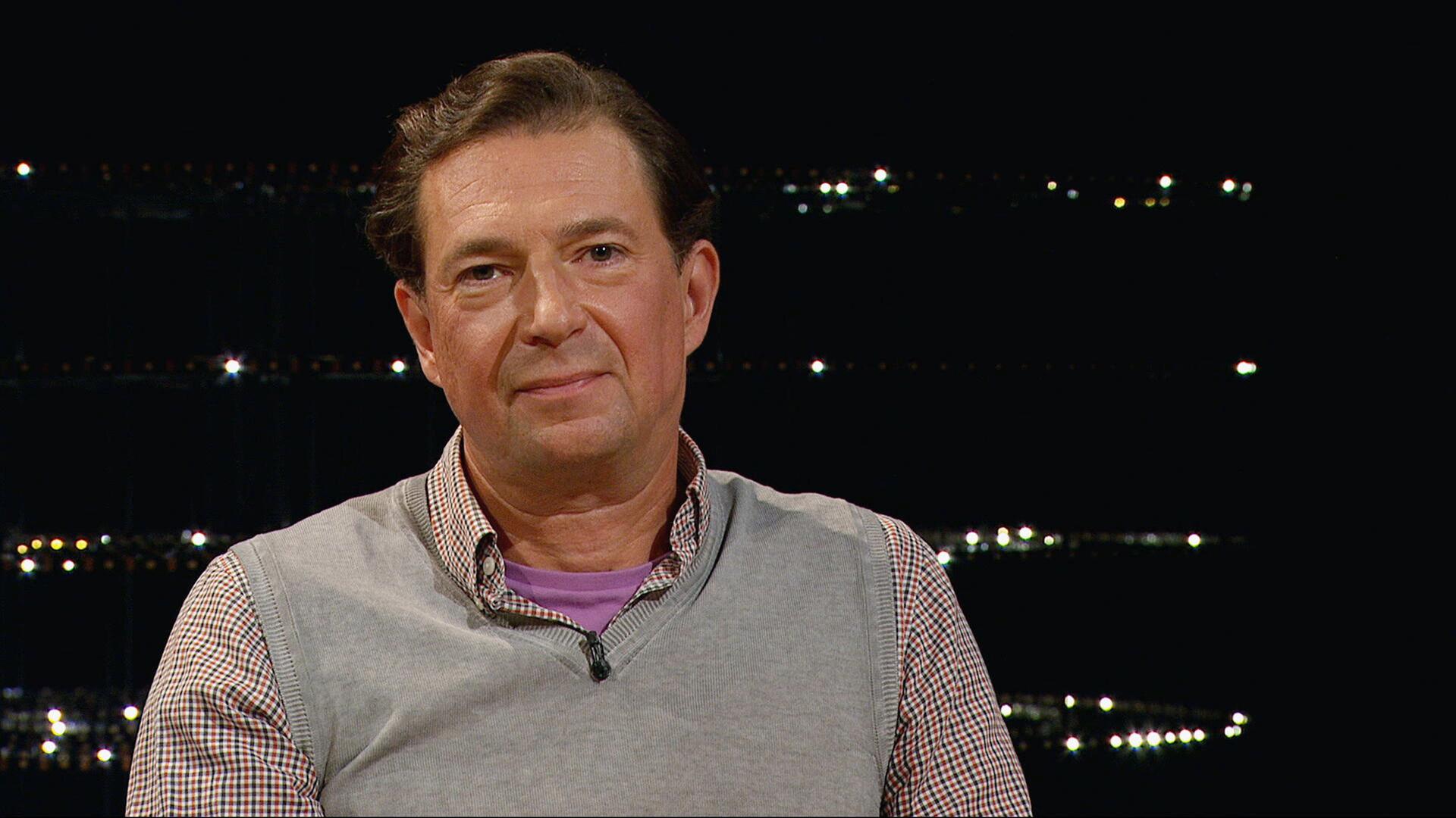 Steve Ayan sitzt in einem Sessel vor dunklem Hintergrund mit verstreuten Lichtern, trägt kariertes Hemd und graue Strickweste und blickt offen und interessiert zur Seite.