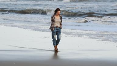 Alice (Julianne Moore) geht in Jeans, T-Shirt und kariertem Hemd an einem einsamen Strand spazieren.