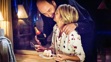 Im gedämpften Licht einer Wohnzimmerlampe beugt sich Thomas Kertez (Kad Merad) mit einer Flasche Sekt in der Hand zu Alice (Laurence Arné), die gerade ein Stück Kuchen auf einem Teller in der Hand hält.