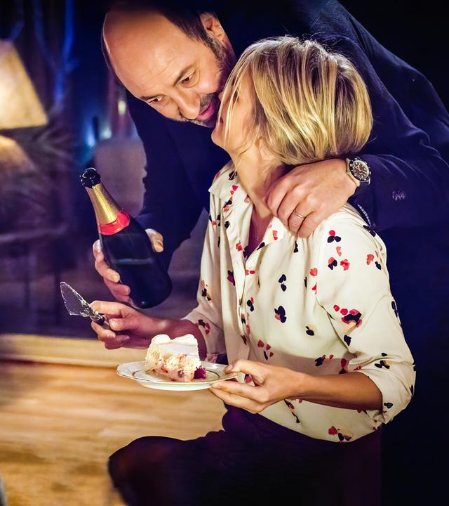 Im gedämpften Licht einer Wohnzimmerlampe beugt sich Thomas Kertez (Kad Merad) mit einer Flasche Sekt in der Hand zu Alice (Laurence Arné), die gerade ein Stück Kuchen auf einem Teller in der Hand hält.