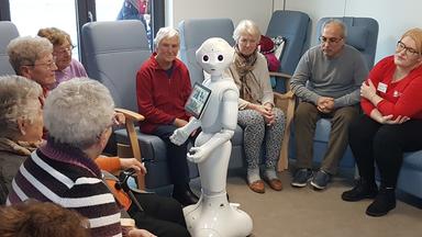 Ein Roboter mit freundlichem Gesicht steht im Raum. Um ihn herum sitzen Menschen und betrachten ihn