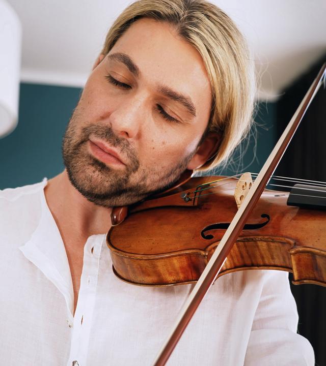 David Garret sitzt in einem Sessel und hält seine Stradivari in seiner Hand und auf seinem Schoß. Er trägt ein lockeres Leinenhemd, auf dem Arm ist deutlich eine Tätowierung sichtbar. Er spricht.
