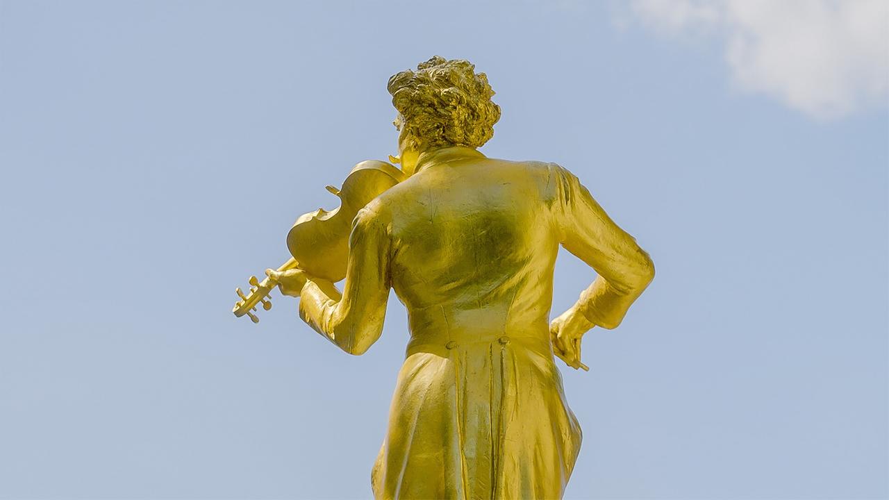 Das Bild zeigt ein goldenes Denkmal, das Johann Strauss darstellt. Die Statue ist von hinten zu sehen und zeigt einen Mann in historischer Kleidung, der eine Violine hält. Seine haarschöpfe sind lockig und voluminös. Die Statue ist vor einem klaren blauen Himmel platziert, was einen guten Kontrast zur goldenen Farbe bietet. Die Kleidung des Mannes ist klassisch und elegant, mit einem langen Gehrock. Im Hintergrund sind keine weiteren erkennbaren Objekte oder Gebäude zu sehen, sodass der Fokus auf der Statue liegt.